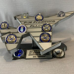 May include: A gray F-16 Fighting Falcon display stand with a silver clock and nine silver challenge coins. The stand has the words "F-16 Fighting Falcon" and "CAPT.  W.R. L. RAY ANTHONY QUARANTE'S TIME 2022 - CHARITY" on it.