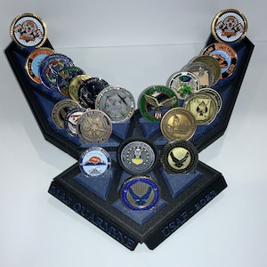 May include: A black, 3D printed display stand shaped like an Air Force wing, with a blue felt lining. The stand holds 20 challenge coins, featuring various designs, including military insignia, flags, and emblems. The stand is labeled "MAJ QUARINE USAF 2023".