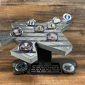 May include: A gray, stylized display stand holding several commemorative coins. The coins have a blue and gold design. A clock and a plaque with text are at the base. The plaque reads "Efia L. Quarymns 151st Logistics Readiness Squadron".