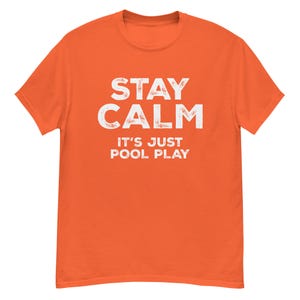 以下が含まれることがあります： オレンジ色のTシャツ。白の文字で「STAY CALM IT'S JUST POOL PLAY」と書かれています。文字はかすれたフォントです。Tシャツは無地で、丸首です。
