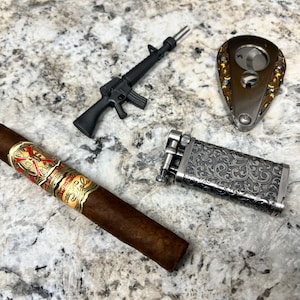 May include: A black toy rifle, a silver cigar cutter, a silver lighter with an engraved floral design, and a brown cigar with a gold and red band that says "Don Francisco" on a gray speckled surface.