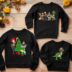 May include: Three black crewneck sweatshirts with different Christmas-themed Toy Story characters. The first sweatshirt features Woody and Buzz decorating a Christmas tree. The second sweatshirt features the Toy Story characters standing together with the text "Merry Christmas". The third sweatshirt features a green dinosaur wearing a Santa hat.