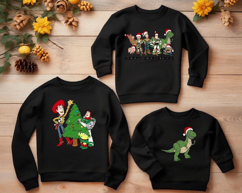 May include: Three black crewneck sweatshirts with different Christmas-themed Toy Story characters. The first sweatshirt features Woody and Buzz decorating a Christmas tree. The second sweatshirt features the Toy Story characters standing together with the text "Merry Christmas". The third sweatshirt features a green dinosaur wearing a Santa hat.