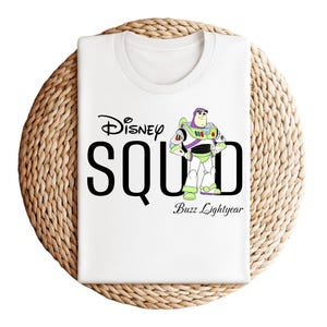 Puede incluir: Camiseta blanca con el texto "Disney SQUAD" en negro y una imagen de dibujos animados de Buzz Lightyear. La camiseta est&aacute; doblada sobre una superficie circular tejida. Las palabras "Buzz Lightyear" est&aacute;n debajo.