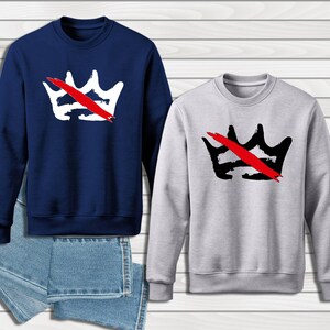 Peut inclure: Deux sweatshirts, un bleu marine et un gris clair, chacun avec un motif de couronne barr&eacute;e d'un trait rouge. Le sweat-shirt bleu marine a une couronne blanche, le gris une couronne noire. Une paire de jeans bleus est visible.