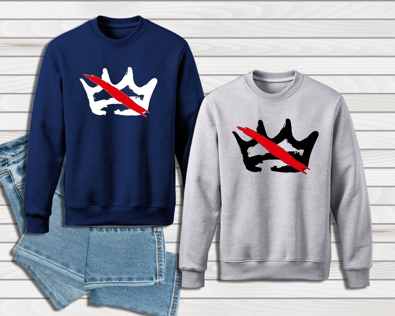 Peut inclure: Deux sweatshirts, un bleu marine et un gris clair, chacun avec un motif de couronne barr&eacute;e d'un trait rouge. Le sweat-shirt bleu marine a une couronne blanche, le gris une couronne noire. Une paire de jeans bleus est visible.