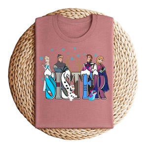 May include: A dusty rose-colored t-shirt featuring the word "SISTER" in a colorful, stylized font. The letters are decorated with cartoon characters from a popular animated film. The shirt is displayed on a woven, circular surface.