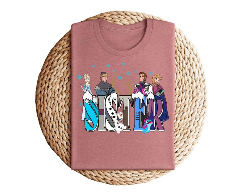 May include: A dusty rose-colored t-shirt featuring the word "SISTER" in a colorful, stylized font. The letters are decorated with cartoon characters from a popular animated film. The shirt is displayed on a woven, circular surface.