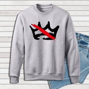 Peut inclure: Un sweat-shirt gris clair avec un motif de couronne noire barr&eacute;e d'une barre rouge. Le sweat-shirt est pr&eacute;sent&eacute; avec un jean en denim bleu. Le motif est centr&eacute; sur le devant.