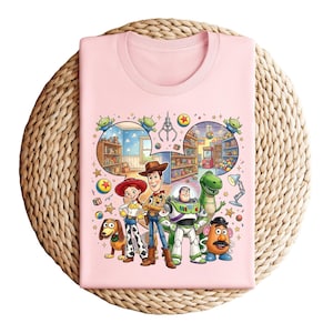 Pode incluir: Uma camiseta rosa claro com um gr&aacute;fico colorido de personagens de um filme de anima&ccedil;&atilde;o. O design inclui Woody, Buzz Lightyear, Jessie, Rex, Sr. Cara de Batata, Slinky Dog e outros elementos do filme. A camiseta est&aacute; dobrada sobre uma superf&iacute;cie tecida.