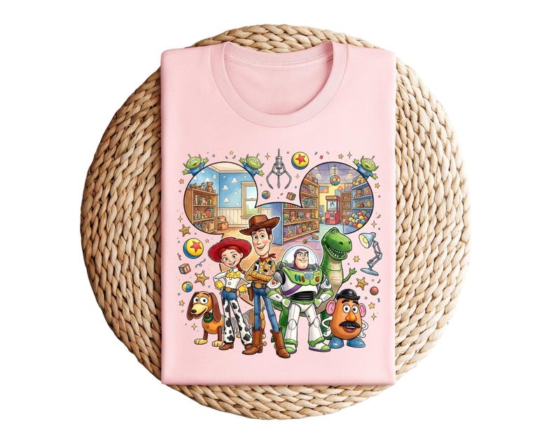 Pode incluir: Uma camiseta rosa claro com um gr&aacute;fico colorido de personagens de um filme de anima&ccedil;&atilde;o. O design inclui Woody, Buzz Lightyear, Jessie, Rex, Sr. Cara de Batata, Slinky Dog e outros elementos do filme. A camiseta est&aacute; dobrada sobre uma superf&iacute;cie tecida.