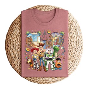 Pode incluir: Camiseta cor de malva com um gr&aacute;fico colorido de personagens de Toy Story. O design inclui Woody, Buzz Lightyear, Jessie, Rex, Sr. Batata e Slinky Dog, com elementos de fundo detalhados.