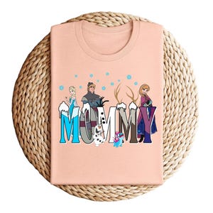 May include: A peach-colored t-shirt featuring the word "MOMMY" in a stylized font with characters from the movie Frozen. The design includes Elsa, Kristoff, Anna, Olaf, and Stitch, with snowflakes and antlers.