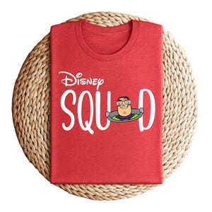Puede incluir: Camiseta roja con el texto blanco "Disney Squad", con una imagen de dibujos animados de Buzz Lightyear. La camiseta est&aacute; doblada cuidadosamente sobre un salvamanteles circular tejido. La camiseta es de cuello redondo.