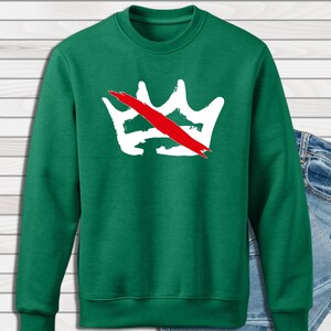 Peut inclure: Un sweat-shirt vert avec un motif de couronne blanche barr&eacute;e d'une bande rouge. Le sweat-shirt est de style col rond et est associ&eacute; &agrave; un jean en denim bleu. Le motif de la couronne est centr&eacute; sur la poitrine.