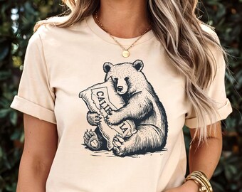 May include: A cream-colored short-sleeve t-shirt featuring a dark navy blue graphic illustration of a bear sitting and holding a pillow. The pillow has a map outline of California and the text CALIFORNIA printed on it in a bold, dark navy blue font.