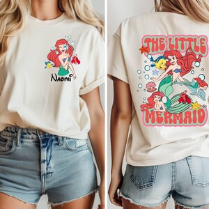 May include: Cream-colored t-shirt featuring a front graphic of a red-haired mermaid with the name "Naomi" and a back graphic that says "The Little Mermaid". The design includes cartoon characters and text.