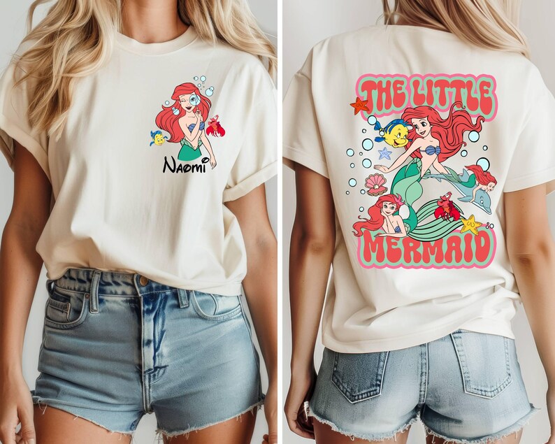 May include: Cream-colored t-shirt featuring a front graphic of a red-haired mermaid with the name "Naomi" and a back graphic that says "The Little Mermaid". The design includes cartoon characters and text.