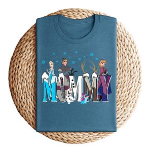 May include: A teal t-shirt featuring the word "MOMMY" in a colorful, stylized font with characters from the movie Frozen. The design includes Elsa, Anna, Kristoff, Olaf, and Sven. The shirt is folded on a woven, circular surface.