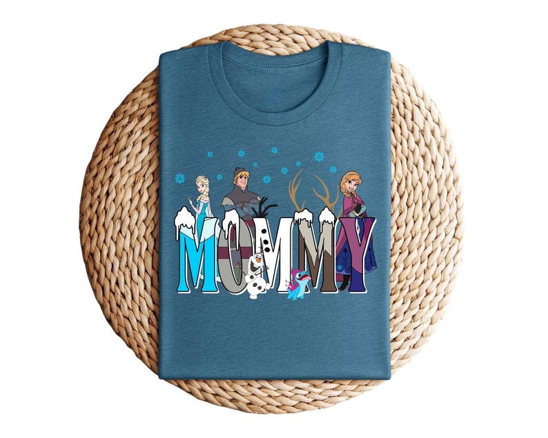 May include: A teal t-shirt featuring the word "MOMMY" in a colorful, stylized font with characters from the movie Frozen. The design includes Elsa, Anna, Kristoff, Olaf, and Sven. The shirt is folded on a woven, circular surface.
