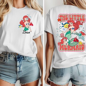 May include: White t-shirt featuring a colorful graphic of a mermaid with red hair, a green tail, and a blue top. The front of the shirt has the name "Linda" in black script. The back of the shirt says "The Little Mermaid" in a retro font.