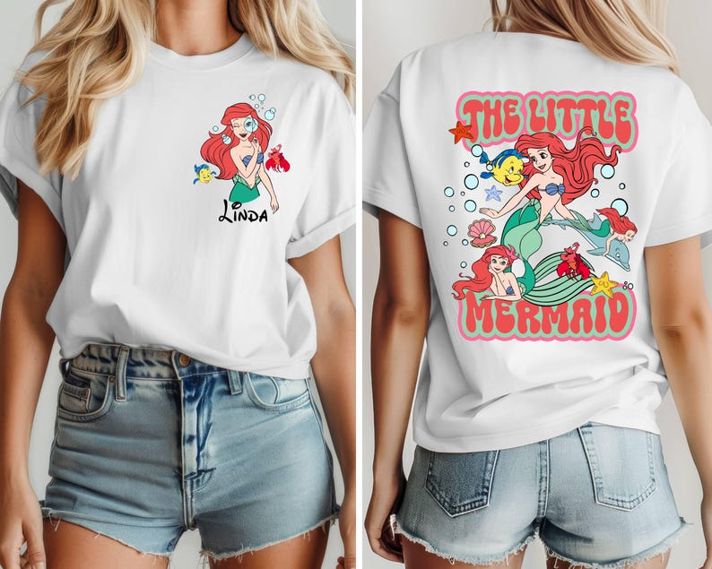May include: White t-shirt featuring a colorful graphic of a mermaid with red hair, a green tail, and a blue top. The front of the shirt has the name "Linda" in black script. The back of the shirt says "The Little Mermaid" in a retro font.