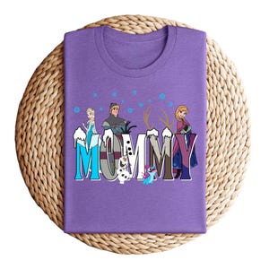 May include: A purple t-shirt with the word "MUMMY" in a stylized font, featuring cartoon characters. The design includes images of Elsa, Anna, Kristoff, Olaf, and Sven from the movie Frozen. The shirt is folded on a woven mat.
