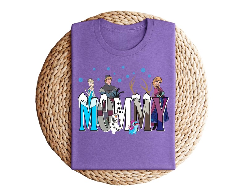 May include: A purple t-shirt with the word "MUMMY" in a stylized font, featuring cartoon characters. The design includes images of Elsa, Anna, Kristoff, Olaf, and Sven from the movie Frozen. The shirt is folded on a woven mat.