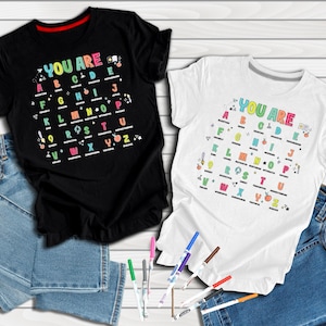 May include: A black t-shirt and a white t-shirt, each featuring a colorful alphabet graphic with the text "YOU ARE" followed by positive adjectives for each letter from A to Z, paired with blue denim jeans and several colorful markers.