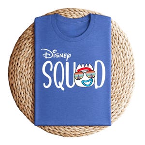 Puede incluir: Una camiseta azul con la frase "Disney Squad" en blanco. La "O" de "Squad" se reemplaza con un personaje de dibujos animados con gafas de sol. La camiseta est&aacute; doblada y colocada sobre una estera circular tejida.