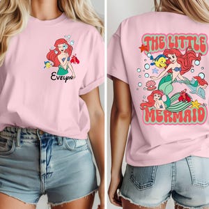 May include: A light pink t-shirt featuring a cartoon illustration of a mermaid with red hair and a green tail. The front of the shirt has the name "Evelyn" below the mermaid. The back of the shirt says "The Little Mermaid" in a retro font.