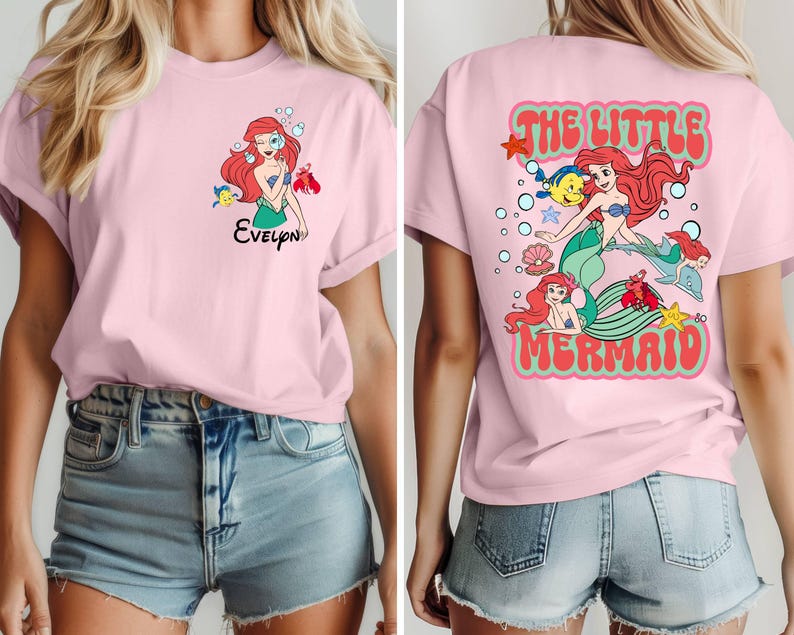 May include: A light pink t-shirt featuring a cartoon illustration of a mermaid with red hair and a green tail. The front of the shirt has the name "Evelyn" below the mermaid. The back of the shirt says "The Little Mermaid" in a retro font.
