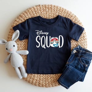 Puede incluir: Camiseta azul marino con la frase "Disney Squad" en blanco. El dise&ntilde;o incluye una cara de dibujos animados con gafas de sol y un gorro rojo. Unos vaqueros azules y un conejito de peluche gris est&aacute;n junto a la camiseta.
