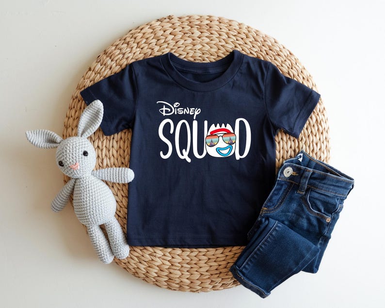 Puede incluir: Camiseta azul marino con la frase "Disney Squad" en blanco. El dise&ntilde;o incluye una cara de dibujos animados con gafas de sol y un gorro rojo. Unos vaqueros azules y un conejito de peluche gris est&aacute;n junto a la camiseta.