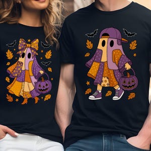 May include: Two black t-shirts featuring graphic prints of ghosts in patchwork-style costumes. One ghost wears a large bow, the other a baseball cap. Both carry purple jack-o'-lantern buckets surrounded by orange autumn leaves and bats.