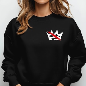 Peut inclure: Sweat-shirt noir avec un motif de couronne blanche barr&eacute;e d'une ligne rouge sur le c&ocirc;t&eacute; gauche de la poitrine. Le sweat-shirt a un col rond et des manches longues. Le motif est un graphisme audacieux.