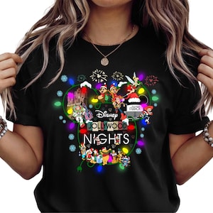 May include: Black t-shirt featuring a colorful graphic design with Disney characters and the text "Disney Jollywood Nights". The design includes images of Mickey Mouse, Minnie Mouse, and other Disney characters, surrounded by Christmas lights.