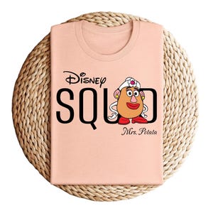 Puede incluir: Camiseta color melocot&oacute;n con el texto negro "Disney SQUAD" y "Mrs. Potato" debajo. La camiseta tiene un gr&aacute;fico de la Sra. Cara de Papa de Toy Story. La camiseta est&aacute; doblada sobre una estera circular tejida.