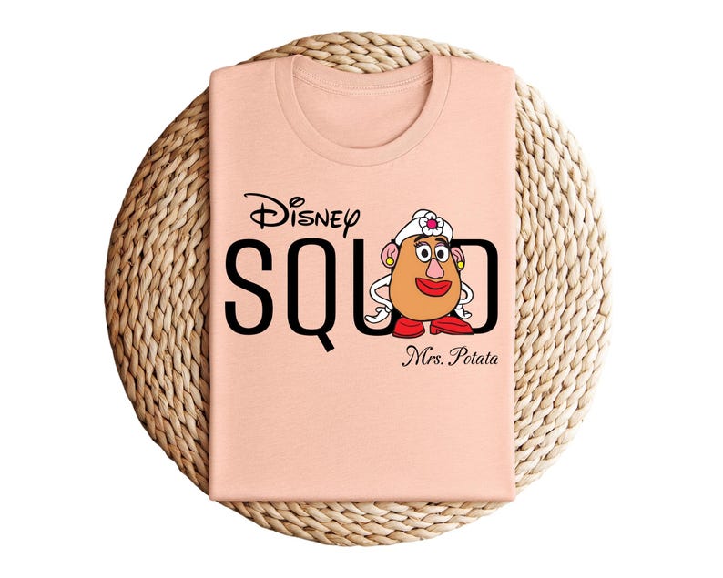 Puede incluir: Camiseta color melocot&oacute;n con el texto negro "Disney SQUAD" y "Mrs. Potato" debajo. La camiseta tiene un gr&aacute;fico de la Sra. Cara de Papa de Toy Story. La camiseta est&aacute; doblada sobre una estera circular tejida.