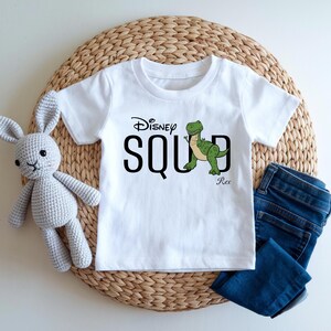 Puede incluir: Camiseta blanca con la frase "Disney Squad" y un dibujo de un dinosaurio verde. La camiseta se muestra con vaqueros azules y un conejo de peluche gris. Los art&iacute;culos est&aacute;n colocados sobre una estera tejida.