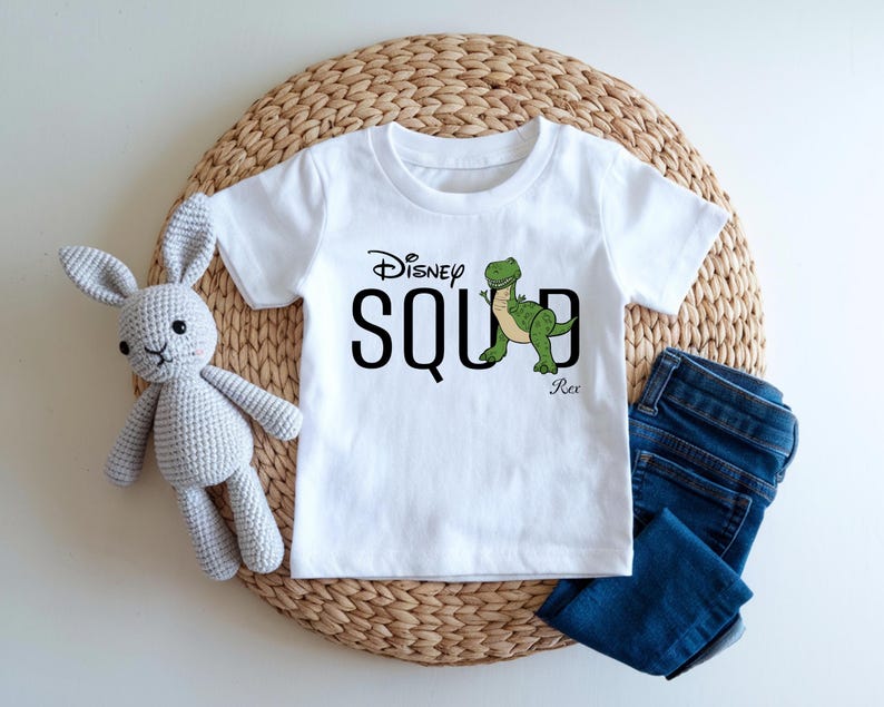 Puede incluir: Camiseta blanca con la frase "Disney Squad" y un dibujo de un dinosaurio verde. La camiseta se muestra con vaqueros azules y un conejo de peluche gris. Los art&iacute;culos est&aacute;n colocados sobre una estera tejida.