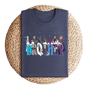 May include: A navy blue t-shirt featuring the word "BROTHER" in a colorful, stylized font. The letters are decorated with cartoon characters and snowflakes. The shirt is folded on a woven, circular mat.