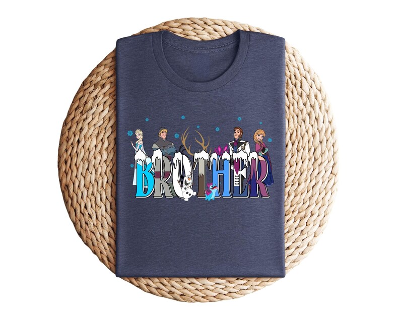 May include: A navy blue t-shirt featuring the word "BROTHER" in a colorful, stylized font. The letters are decorated with cartoon characters and snowflakes. The shirt is folded on a woven, circular mat.