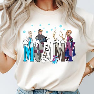 May include: Cream-colored t-shirt featuring the word "MOMMY" in a stylized font with cartoon characters from the movie Frozen. The letters are decorated with snow and the characters include Elsa, Anna, Kristoff, Olaf, and Sven.