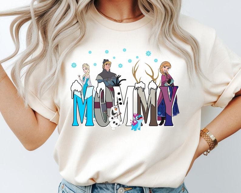 May include: Cream-colored t-shirt featuring the word "MOMMY" in a stylized font with cartoon characters from the movie Frozen. The letters are decorated with snow and the characters include Elsa, Anna, Kristoff, Olaf, and Sven.