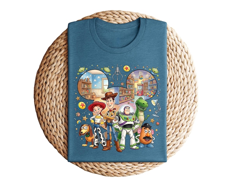 Pode incluir: Camiseta azul-petr&oacute;leo com um gr&aacute;fico colorido de personagens do filme Toy Story. O design inclui Woody, Buzz Lightyear, Jessie, Rex, Sr. Batata e Slinky Dog, com um fundo de loja de brinquedos.