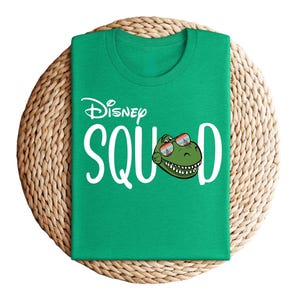 Puede incluir: Una camiseta verde con la frase "Disney Squad" en blanco. La letra "O" de "Squad" es reemplazada por una cabeza de dinosaurio de dibujos animados con gafas de sol. La camiseta est&aacute; sobre un salvamanteles redondo tejido.