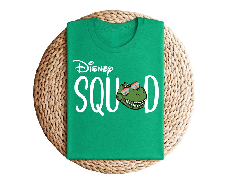 Puede incluir: Una camiseta verde con la frase "Disney Squad" en blanco. La letra "O" de "Squad" es reemplazada por una cabeza de dinosaurio de dibujos animados con gafas de sol. La camiseta est&aacute; sobre un salvamanteles redondo tejido.