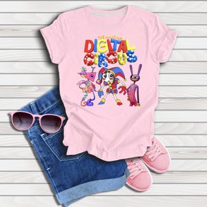 May include: A light pink t-shirt featuring a graphic print of The Amazing Digital Circus with three animated characters, blue denim shorts, pink sunglasses, and pink sneakers. The shirt displays the colorful text The Amazing Digital Circus.