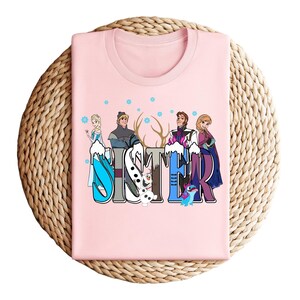 May include: A light pink t-shirt featuring the word "SISTER" in a colorful, winter-themed design. The design includes cartoon characters and snowflakes. The shirt is folded on a woven, circular surface.